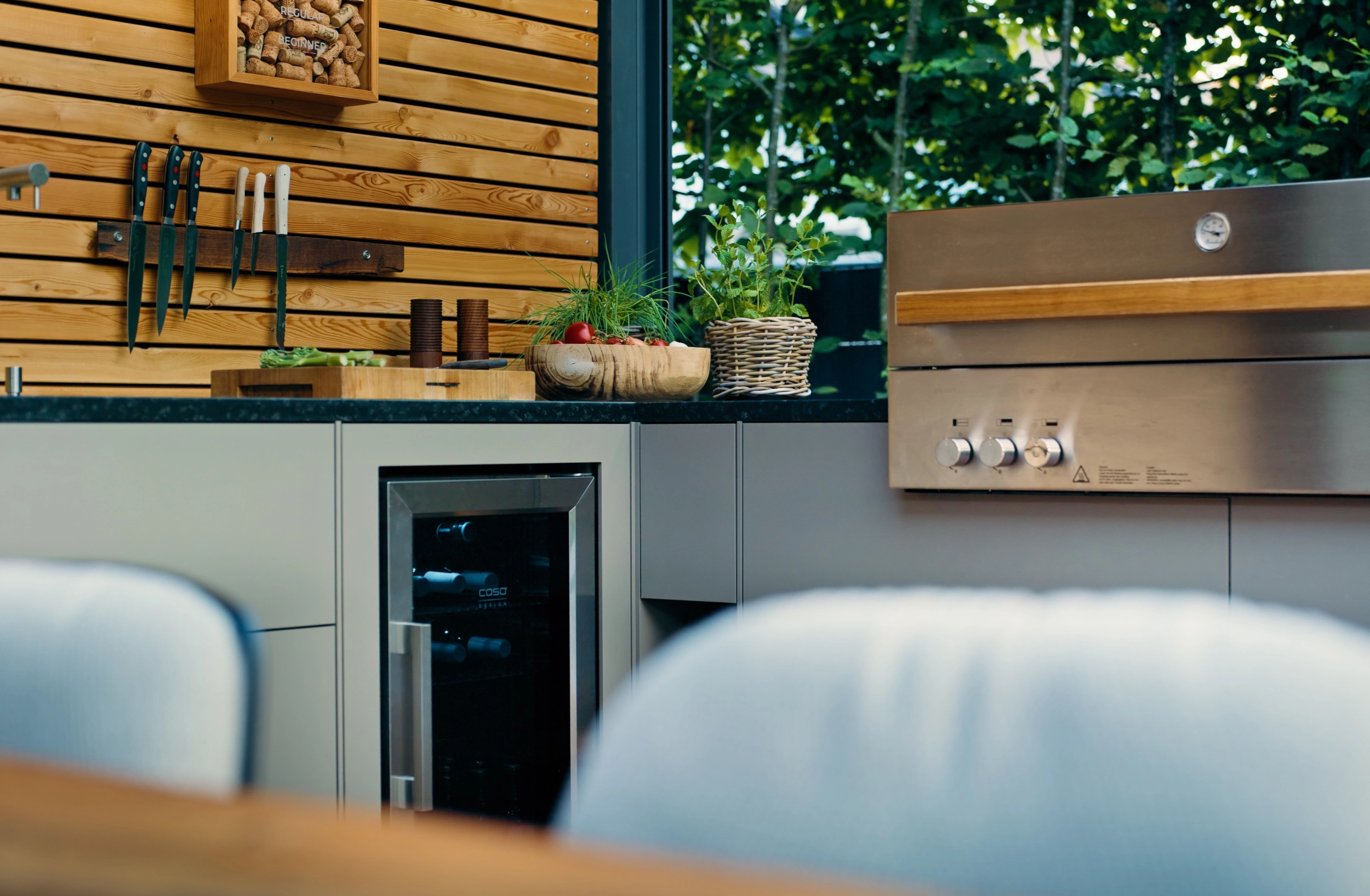 Außenküche mit Weinkühlschrank und Gasgrill vor einer Holzwand, Kräuter auf der Arbeitsplatte