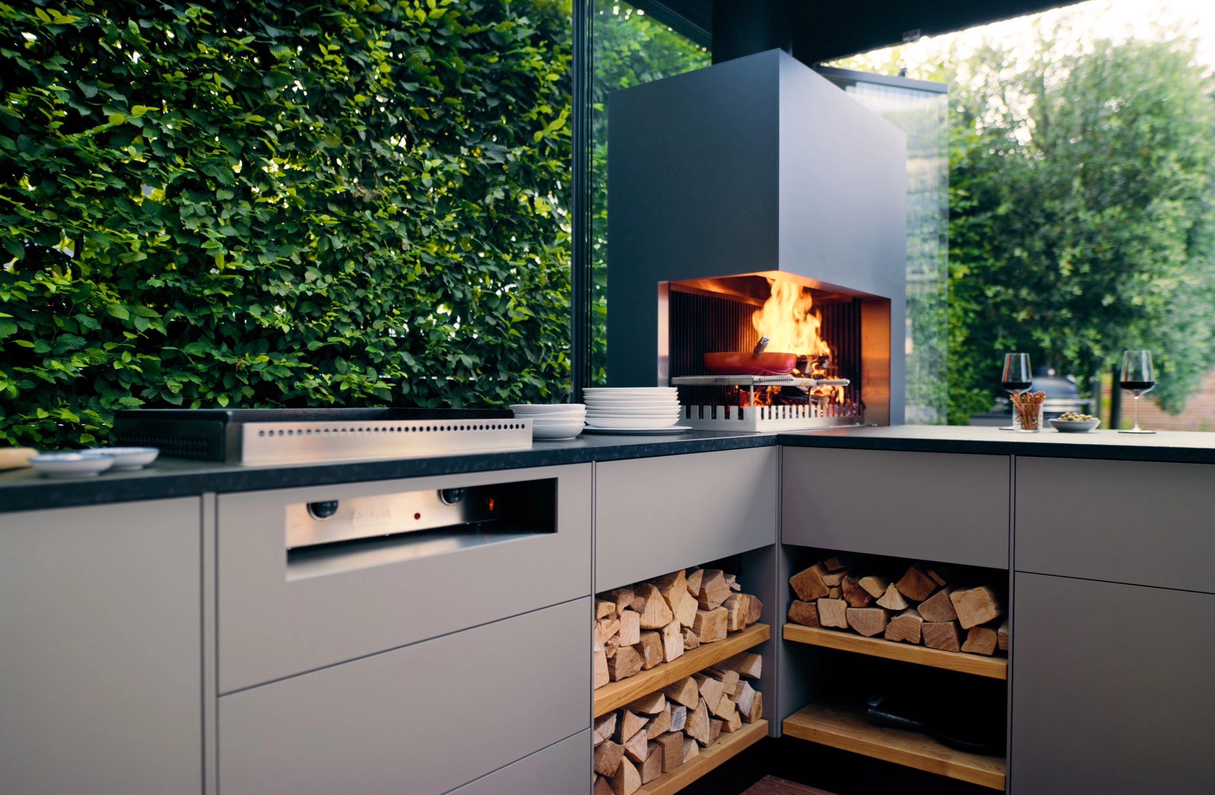 Überdachte Ambienteküche in L-Form mit offenem Feuer, Plancha und Holzlager vor grüner Hecke