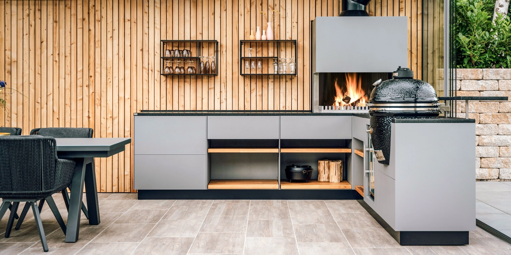 Graue Outdoorküche mit Grill-Feuerstelle und Kamado vor Holzlattenwand, davor der Esstisch