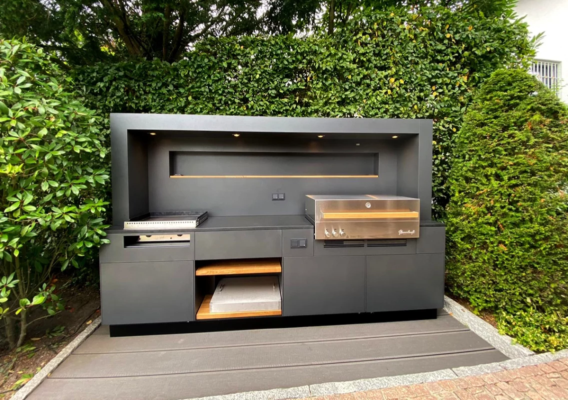 Anthrazitfarbene Outdoorküche mit Grill und Pizzaofen vor hoher Hecke
