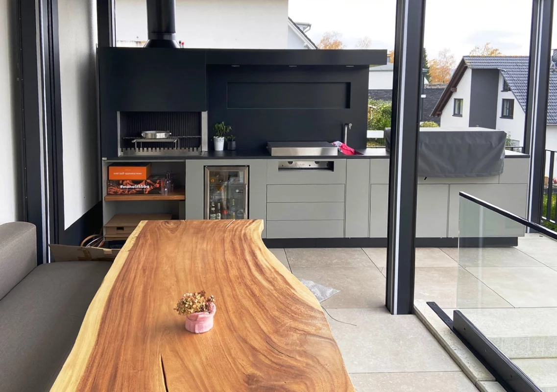 Überdachte Außenküche mit Grill, davor ein Esstisch aus Massivholz